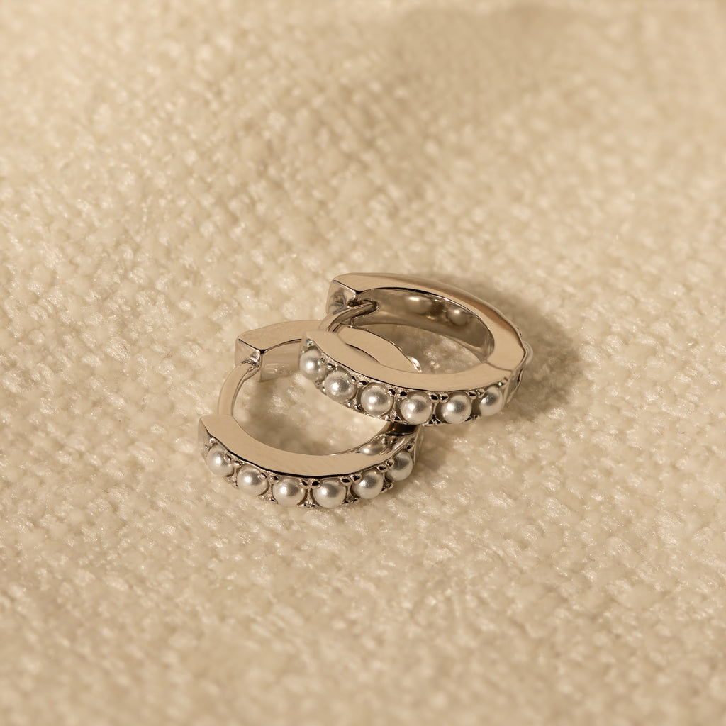 A pair of silver-toned huggie earrings with pearl accents lying flat on a soft beige textile surface.
