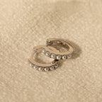 A pair of silver-toned huggie earrings with pearl accents lying flat on a soft beige textile surface.