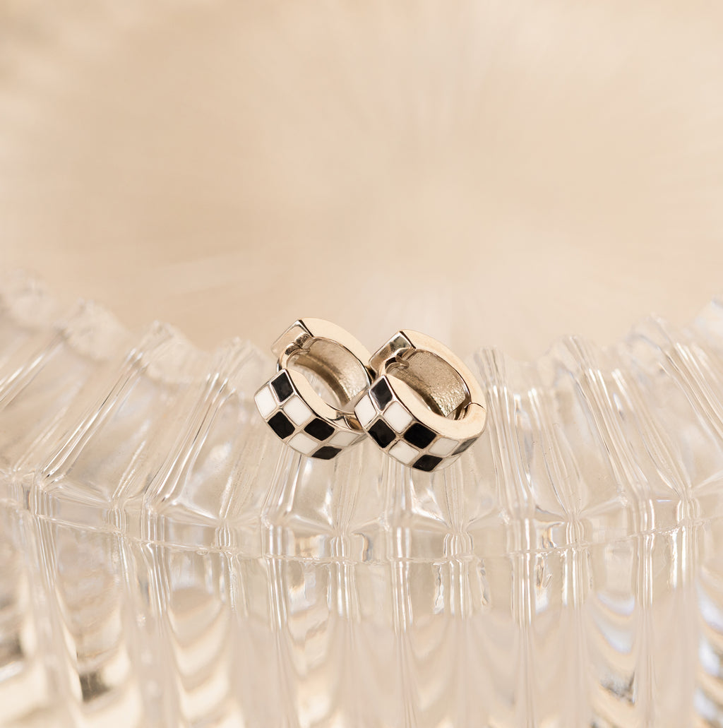 Add flair to any look with our stylish Checkered Huggies—silver hoop earrings featuring a bold black and white checker pattern. Displayed elegantly on clear textured glass.