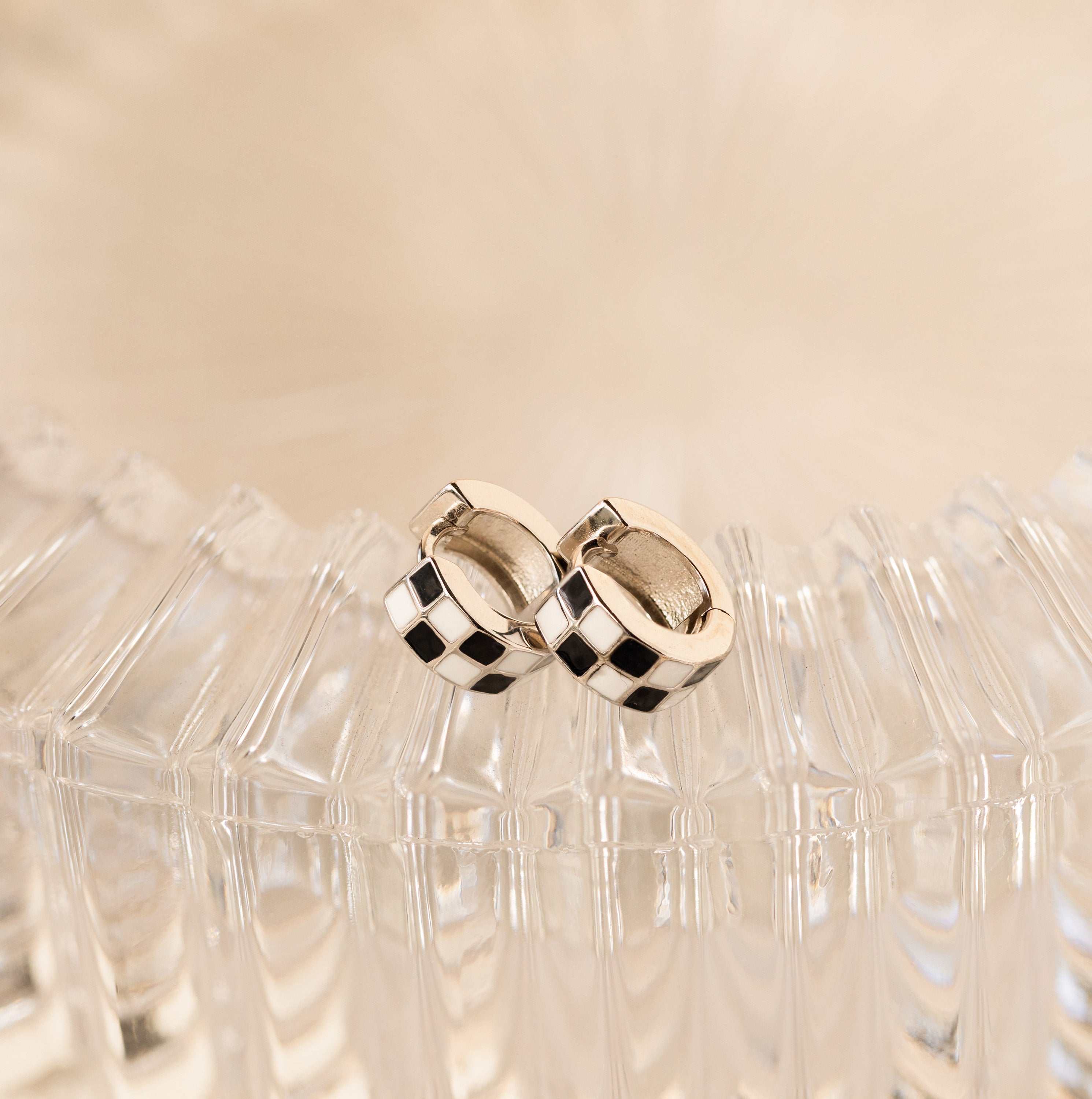 Add flair to any look with our stylish Checkered Huggies—silver hoop earrings featuring a bold black and white checker pattern. Displayed elegantly on clear textured glass.