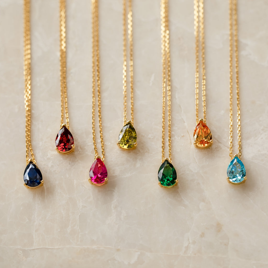 Seven Ophelia Birthstone Necklaces, each featuring a gold teardrop gemstone in a different color, are arranged in a row on marble—ideal for creating a personalized minimalist birthstone necklace set.