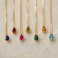 Seven Ophelia Birthstone Necklaces, each featuring a gold teardrop gemstone in a different color, are arranged in a row on marble—ideal for creating a personalized minimalist birthstone necklace set.