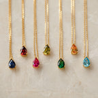 Seven Ophelia Birthstone Necklaces, each featuring a gold teardrop gemstone in a different color, are arranged in a row on marble—ideal for creating a personalized minimalist birthstone necklace set.