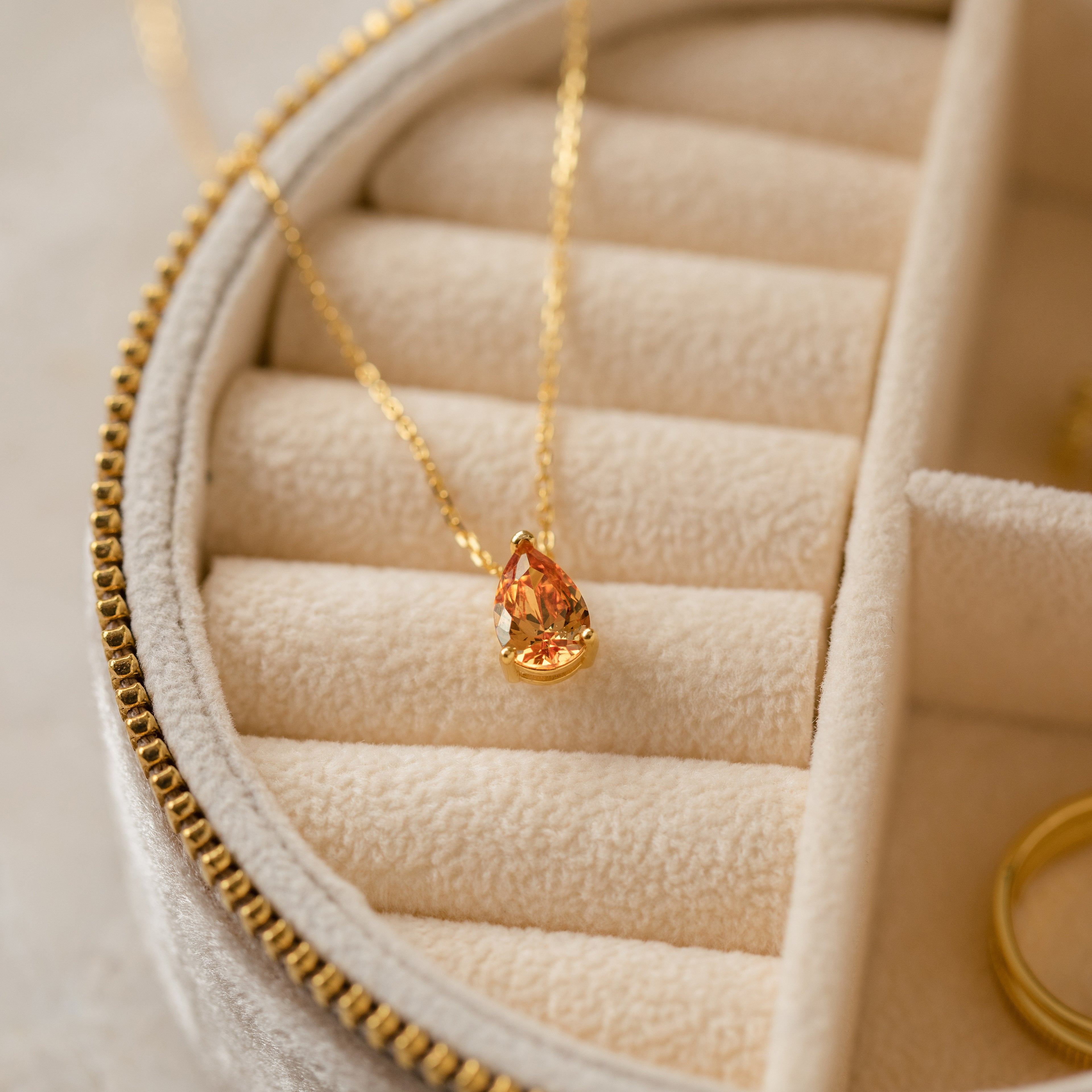 Ophelia Birthstone Necklace: A gold minimalist necklace with a teardrop-shaped orange birthstone pendant, presented in a cream velvet jewelry box.