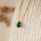 The Ophelia Birthstone Necklace features a teardrop-shaped green gemstone pendant, elegantly displayed on a textured beige surface.