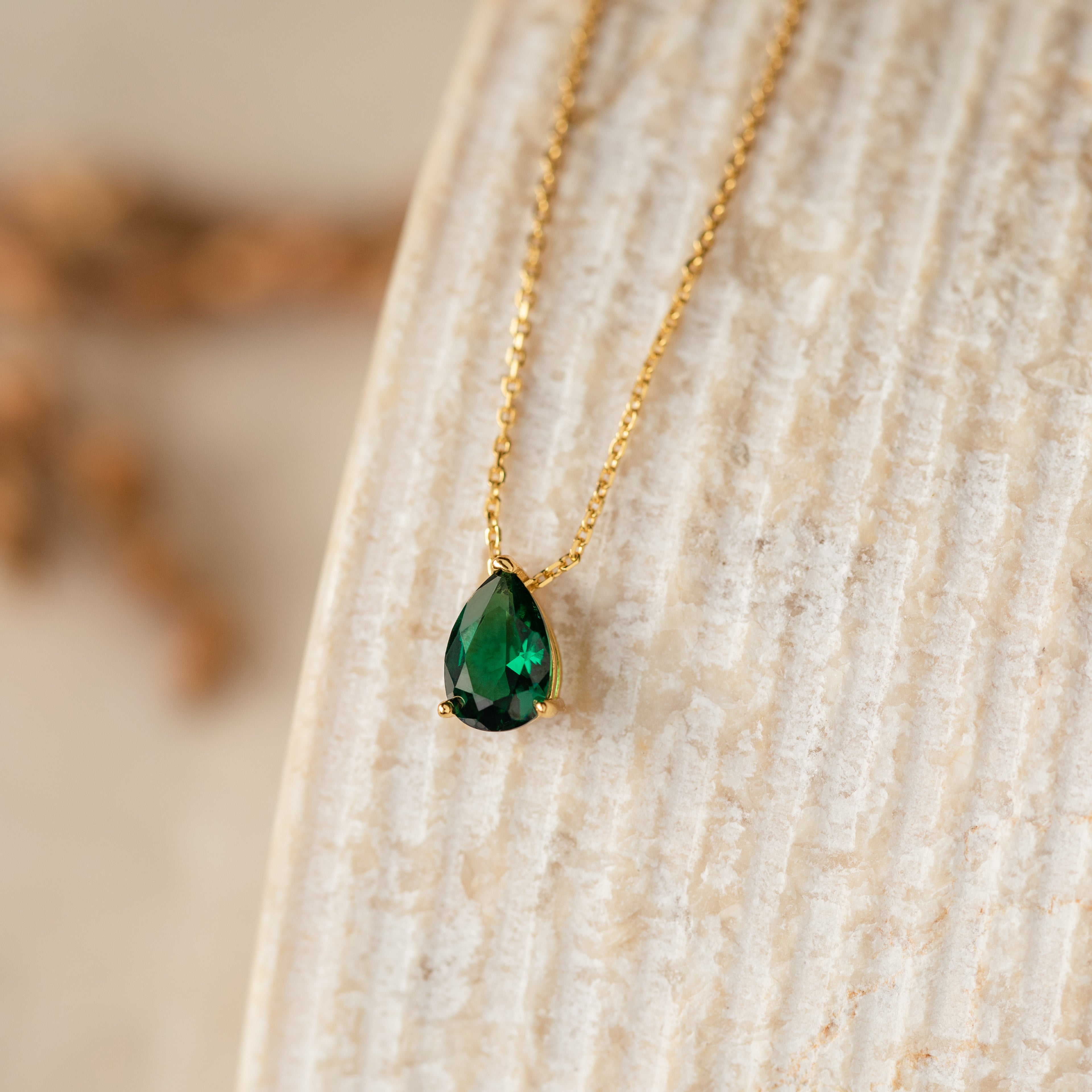 The Ophelia Birthstone Necklace features a teardrop-shaped green gemstone pendant, elegantly displayed on a textured beige surface.