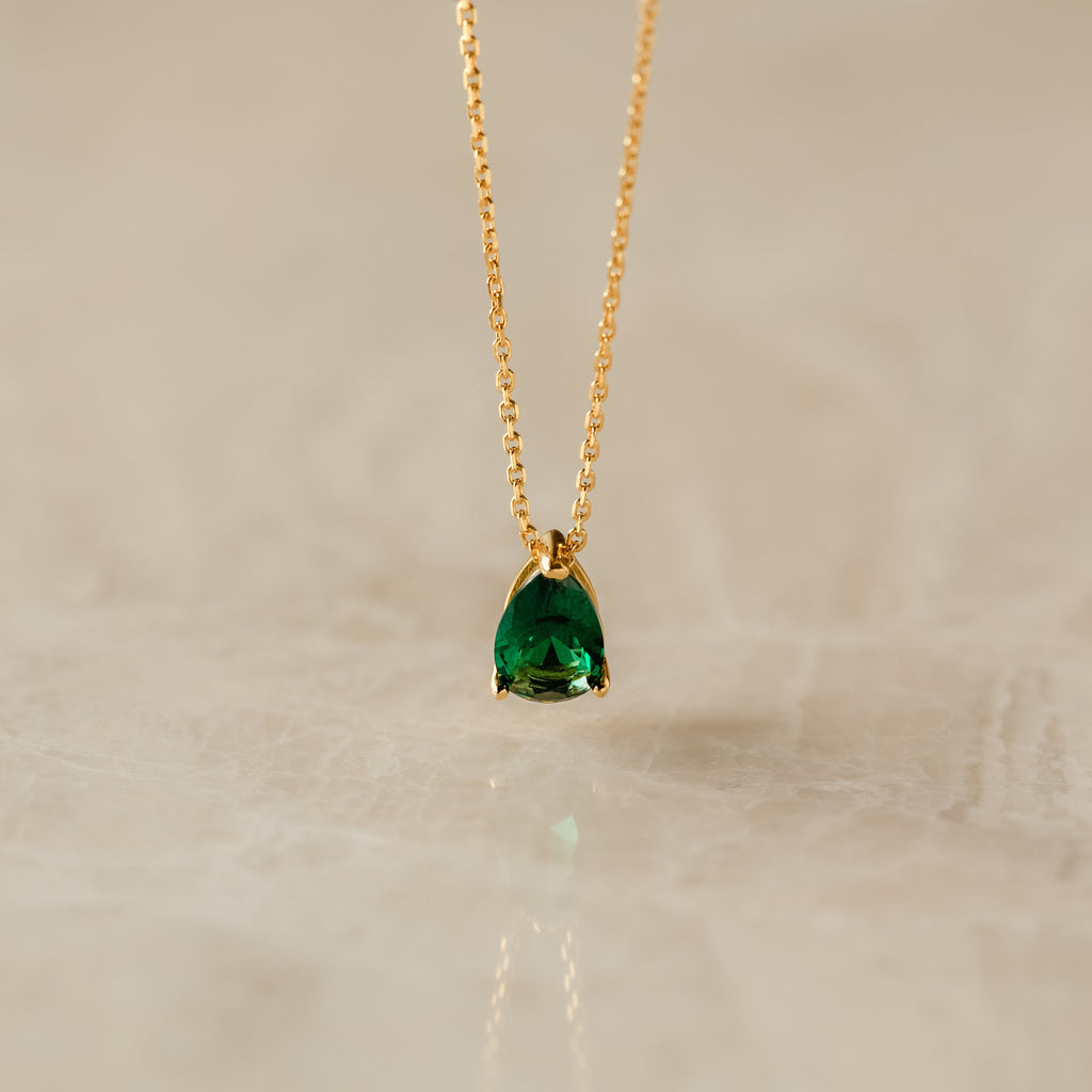 Ophelia Birthstone Necklace with a teardrop green gemstone pendant on a gold chain, elegantly displayed on a light marble surface.
