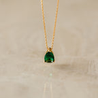 Ophelia Birthstone Necklace with a teardrop green gemstone pendant on a gold chain, elegantly displayed on a light marble surface.
