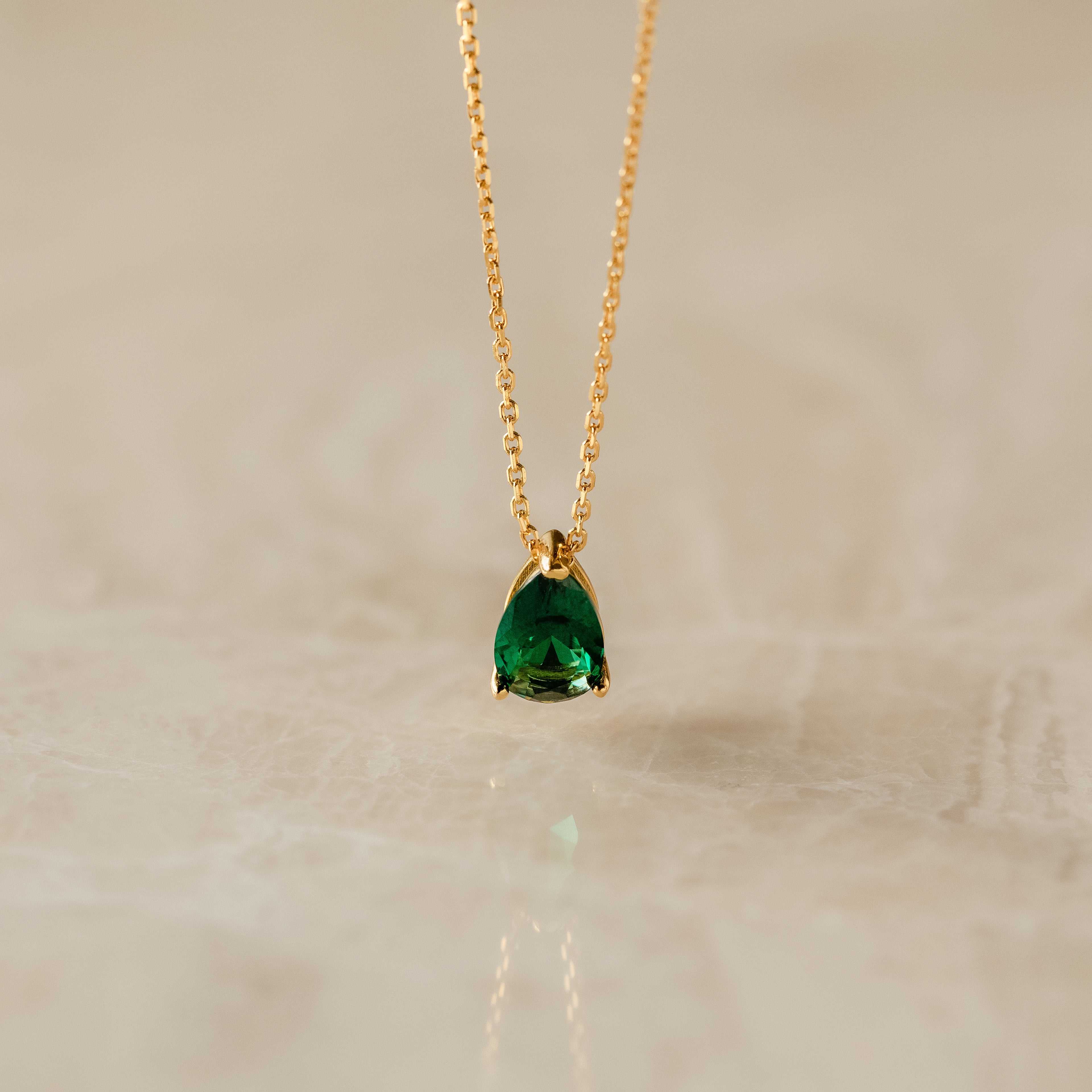 Ophelia Birthstone Necklace with a teardrop green gemstone pendant on a gold chain, elegantly displayed on a light marble surface.