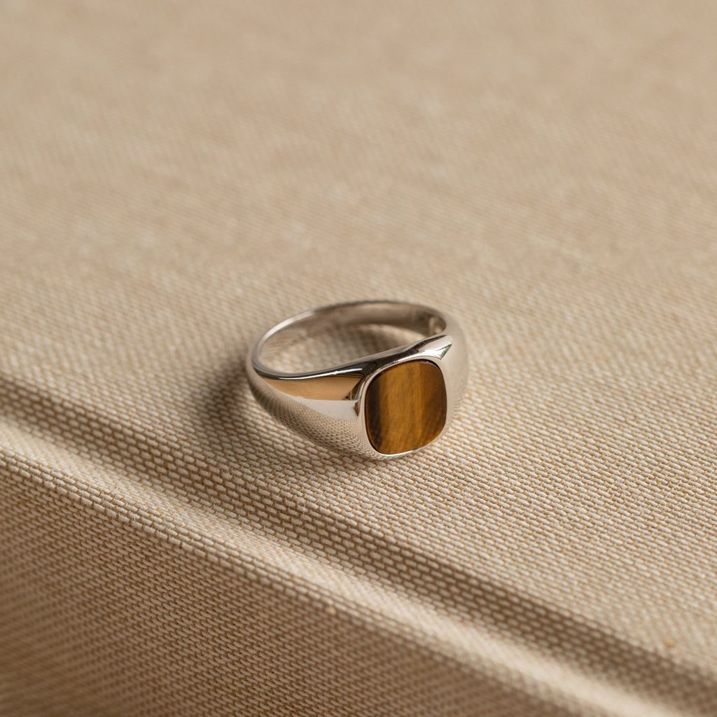 The Tigers Eye Signet Ring in Sterling Silver features a bold oval brown stone, making a statement as it sits atop a textured beige fabric-covered book.