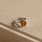 The Tigers Eye Signet Ring in Sterling Silver features a bold oval brown stone, making a statement as it sits atop a textured beige fabric-covered book.