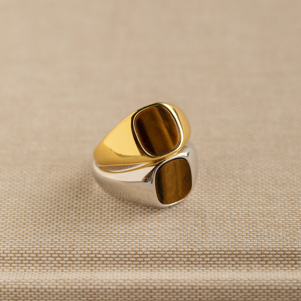 Two Tigers Eye Signet Rings—one gold, one silver—feature striking brown stone insets on a beige fabric background. Each bold piece is perfect for elevating your look.