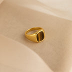 The Tigers Eye Signet Ring in 18K Gold, featuring a brown rectangular stone, rests on a soft beige fabric background.