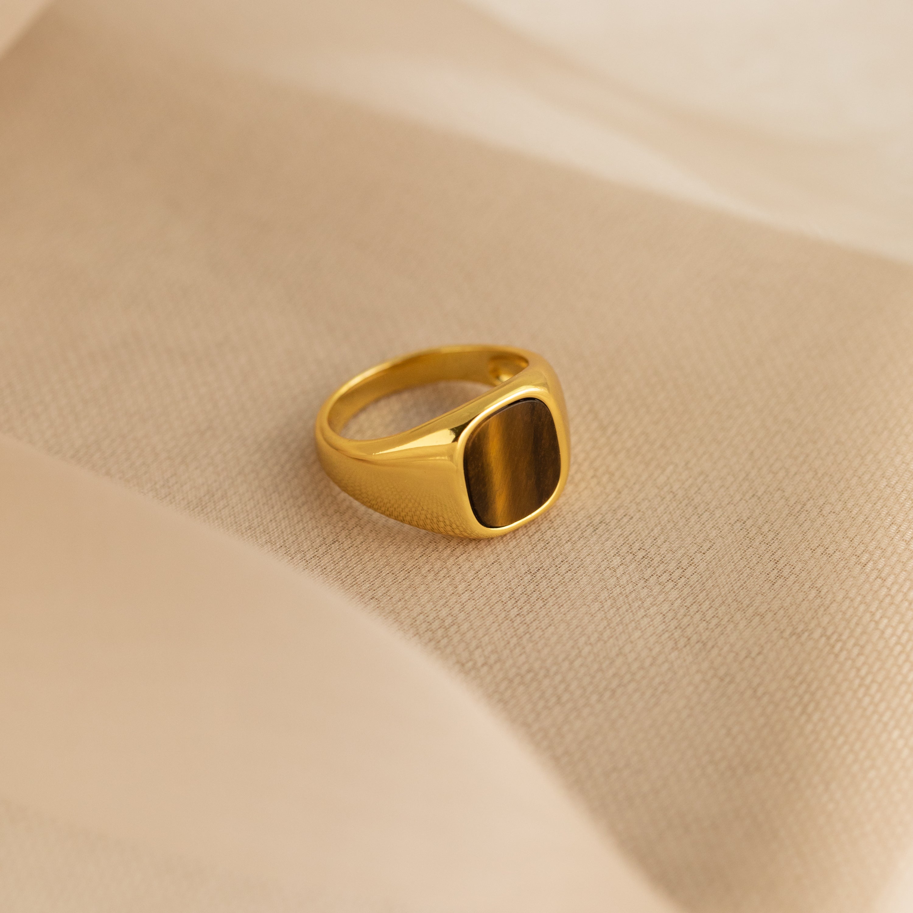 The Tigers Eye Signet Ring in 18K Gold, featuring a brown rectangular stone, rests on a soft beige fabric background.
