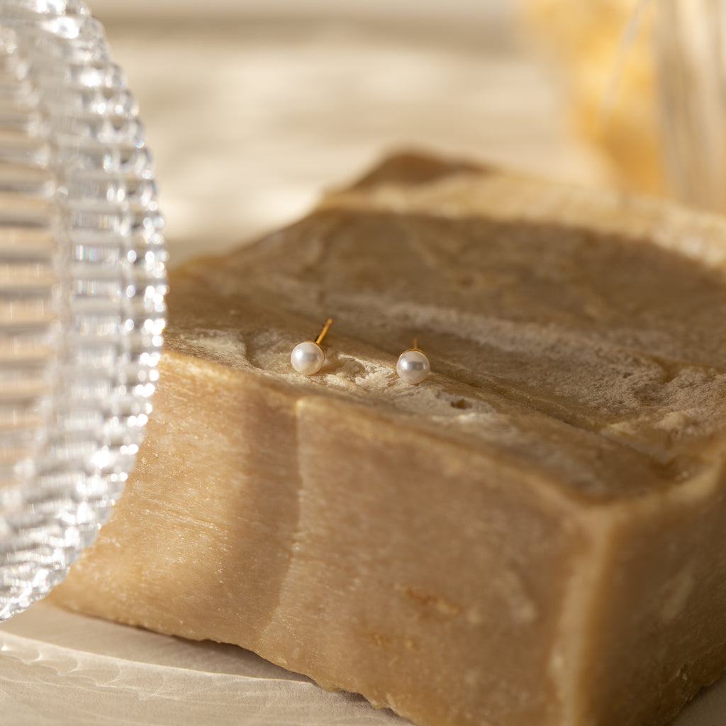 The Dainty Pearl Studs are displayed on a textured stone surface with soft, warm lighting, highlighting their timeless appeal as elegant minimalist earrings.