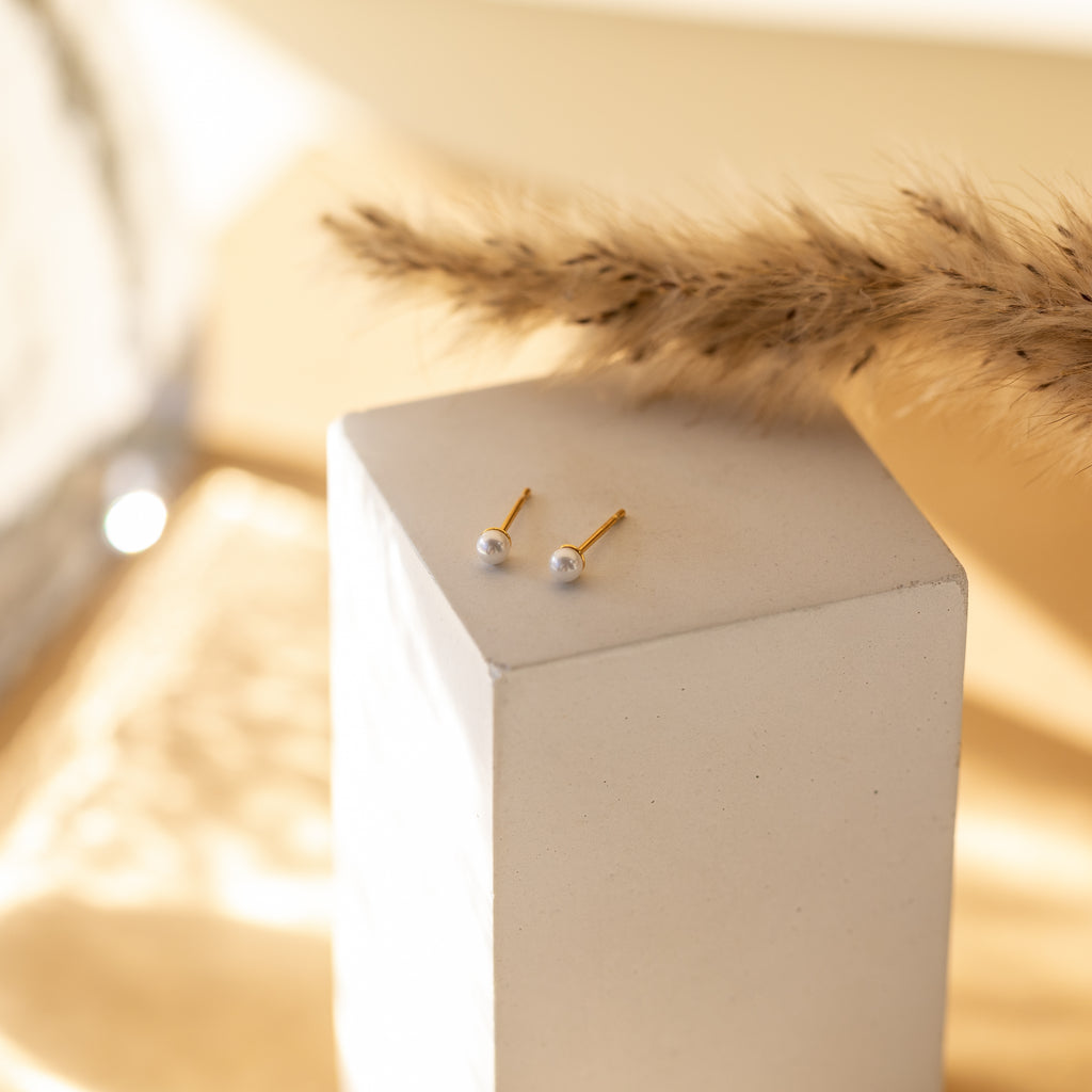 The Dainty Pearl Studs in 18K Gold rest on a white cube with dried grass behind them—perfect minimalist earrings for easy, effortless stacking.
