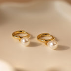 The Dainty Pearl Drop Huggies in 18K Gold showcase delicate white pearl drops on gold hoops, resting on a smooth cream surface—an elegant earring choice.