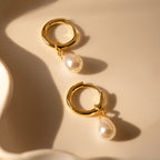 The Dainty Pearl Drop Huggies in 18K Gold feature gold hoops with teardrop pearls, displayed on a cream backdrop in soft lighting—elegant earrings ideal for any occasion.