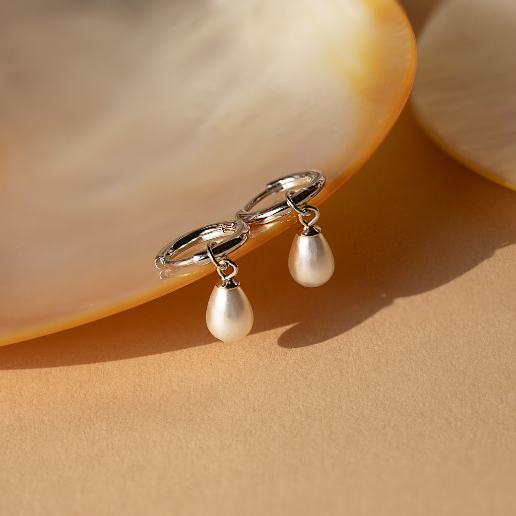 Dainty Pearl Drop Huggies in Sterling Silver—elegant silver hoop earrings with dangling pearls, showcased against a shell on a beige background.