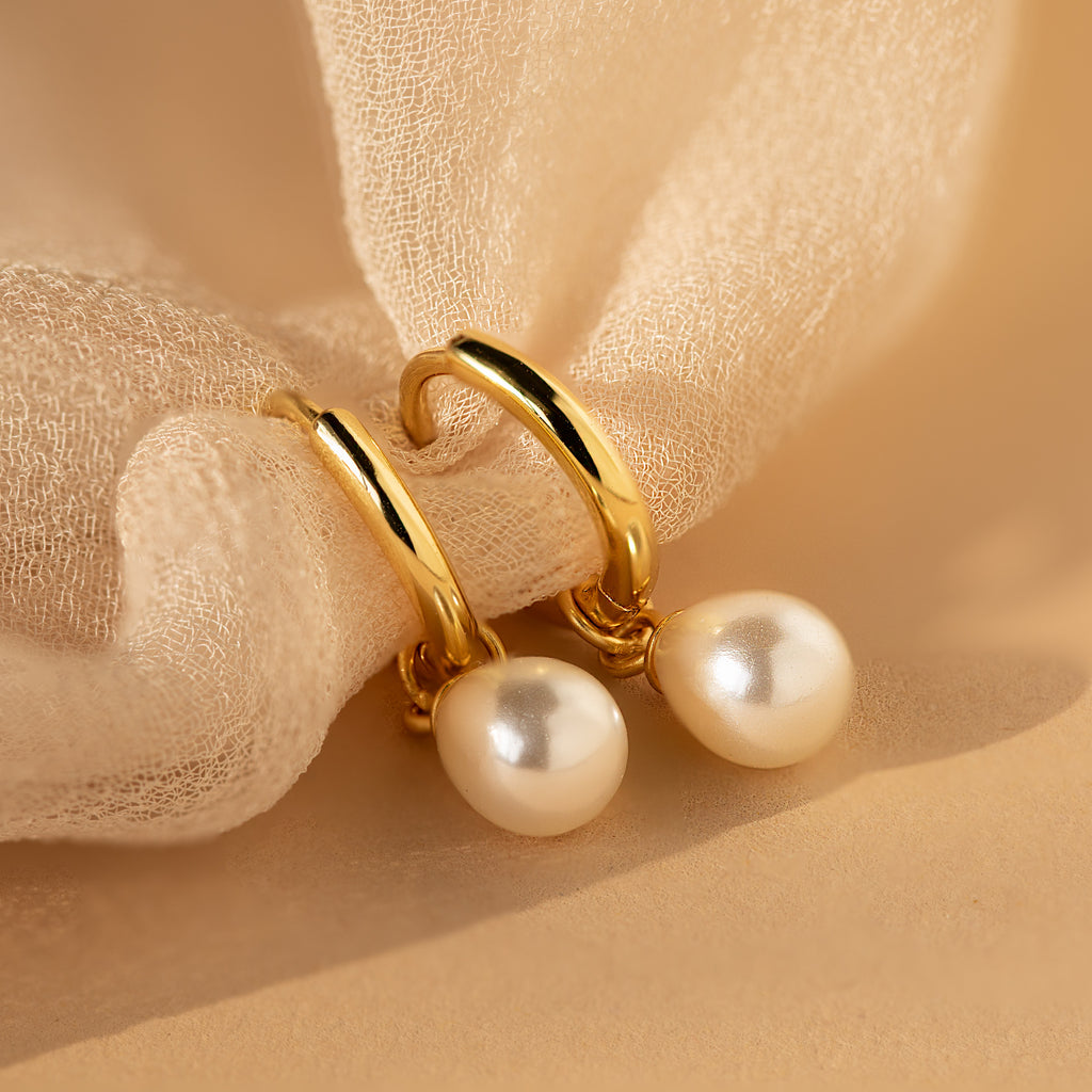 The Dainty Pearl Drop Huggies in 18K Gold feature elegant gold hoops with dangling white pearls, beautifully displayed on soft beige fabric.