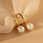 The Dainty Pearl Drop Huggies in 18K Gold feature elegant gold hoops with dangling white pearls, beautifully displayed on soft beige fabric.