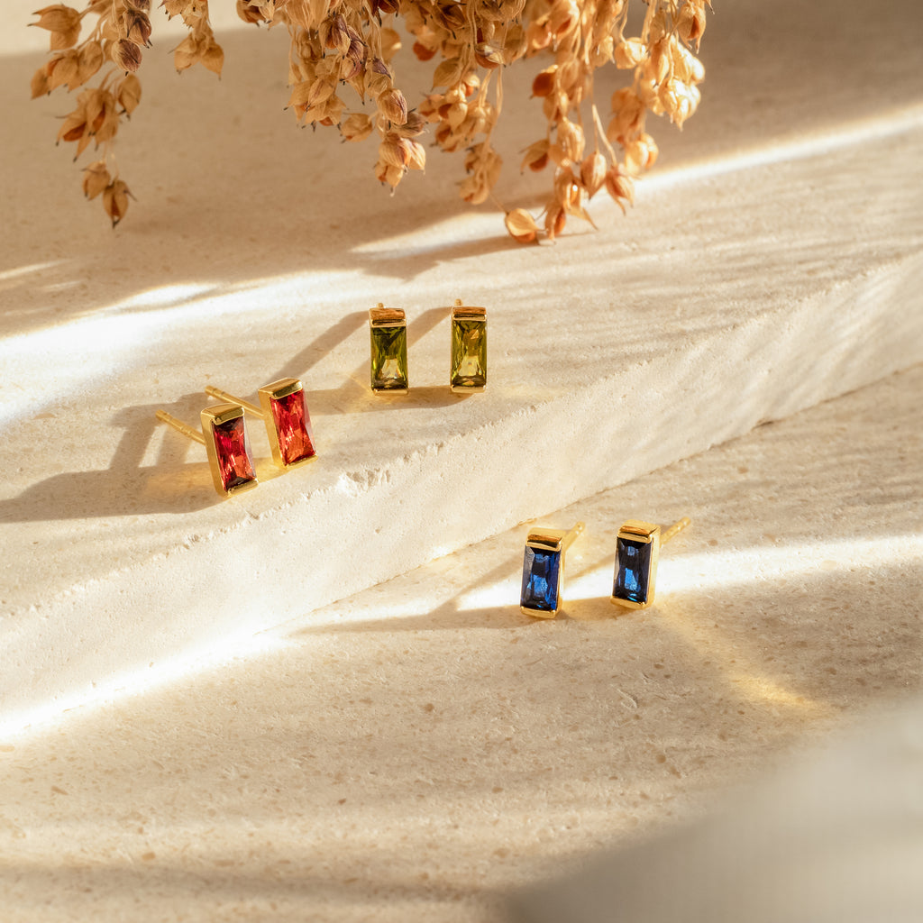 Three pairs of Baguette Birthstone Studs in gold, each featuring red, green, and blue gems, are displayed on a textured cream surface—ideal as birthstone earrings or personalized gifts.