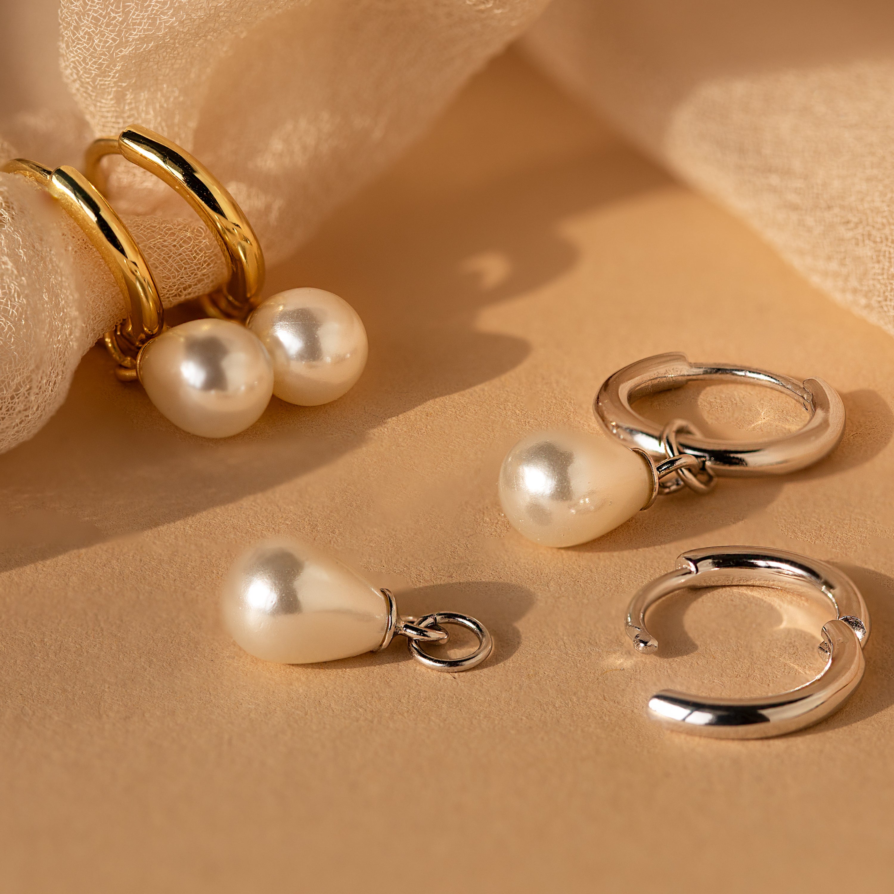 The Dainty Pearl Drop Huggies in Sterling Silver rest gracefully on a soft beige fabric background, showcasing elegant hoops with delicate dangling pearls.
