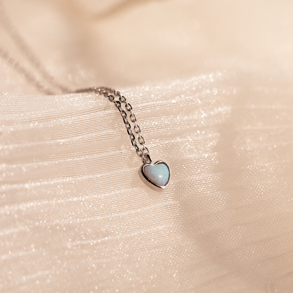 A Tiny Opal Heart Necklace with a delicate heart-shaped pendant rests on textured, cream-colored fabric.