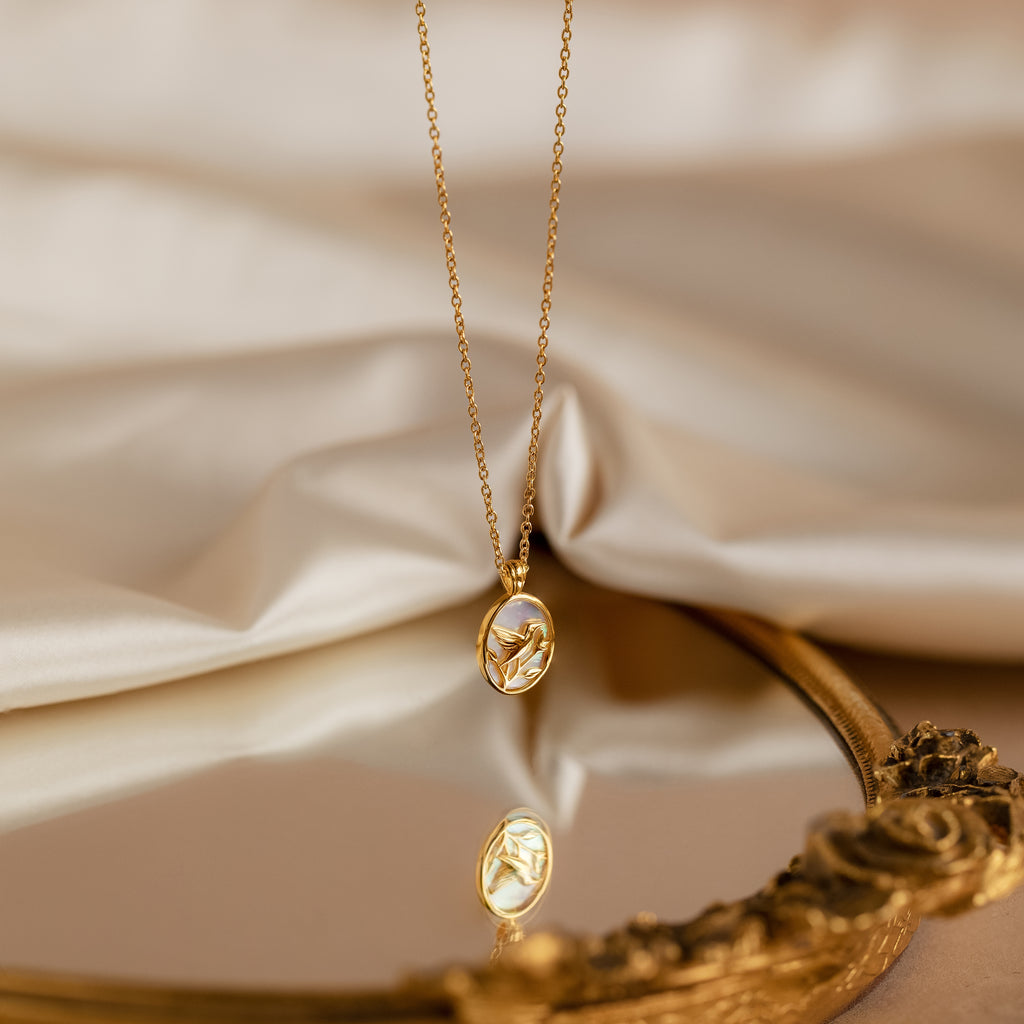 Pearl Hummingbird Necklace in 18K Gold rests on fabric, reflected in an ornate mirror—a vintage-inspired piece that adds elegant simplicity to any look.