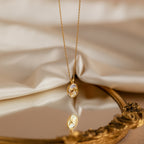 Pearl Hummingbird Necklace in 18K Gold rests on fabric, reflected in an ornate mirror—a vintage-inspired piece that adds elegant simplicity to any look.