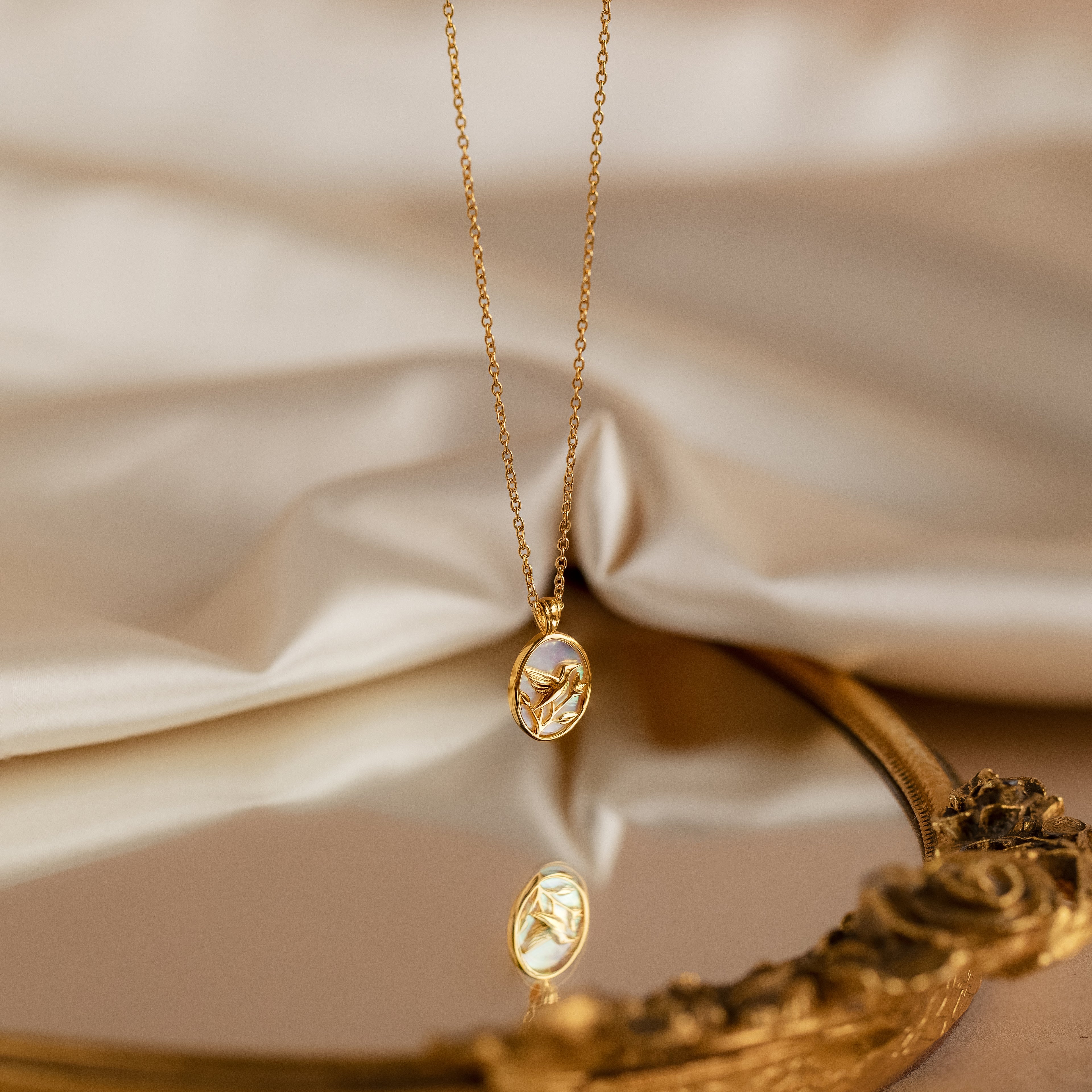 Pearl Hummingbird Necklace in 18K Gold rests on fabric, reflected in an ornate mirror—a vintage-inspired piece that adds elegant simplicity to any look.