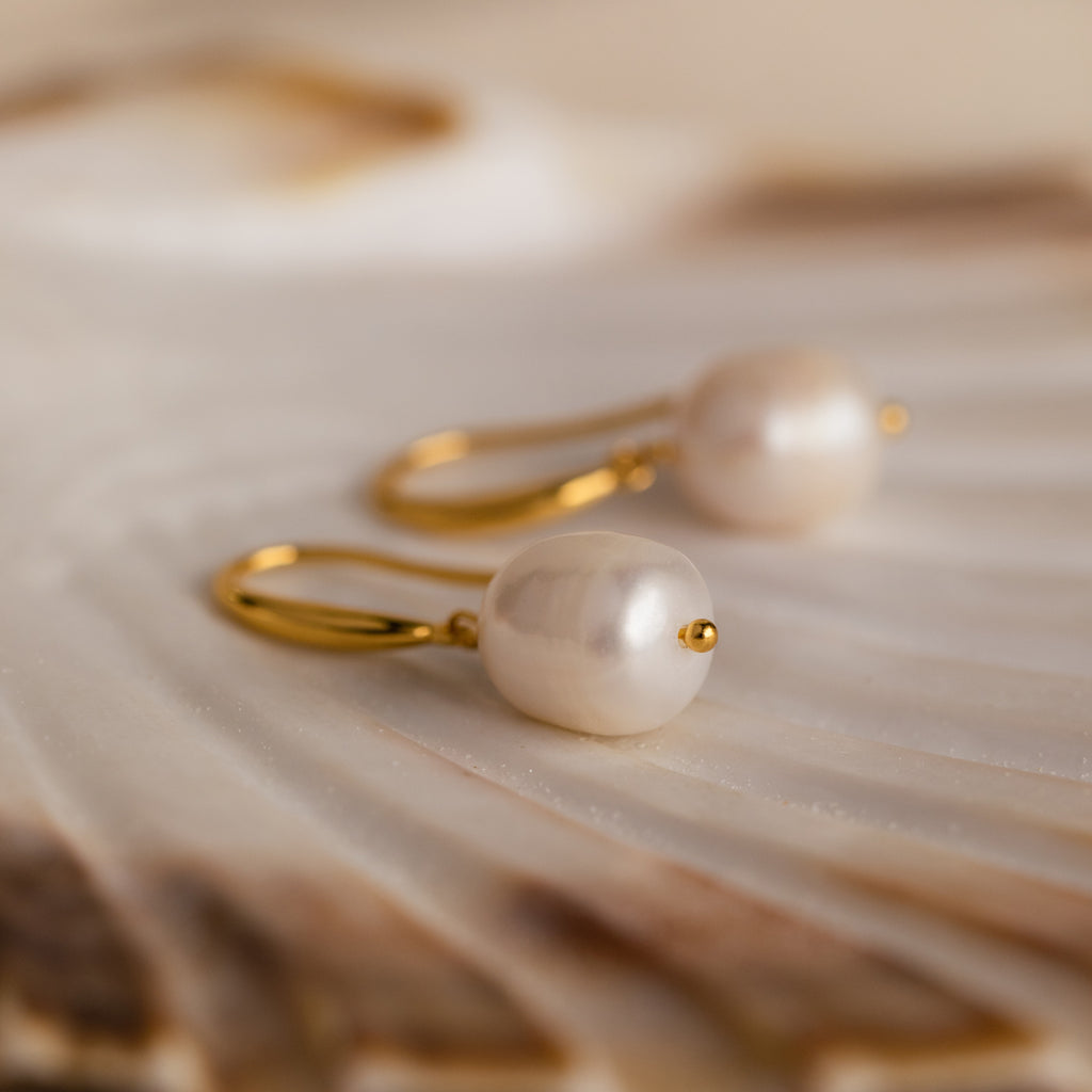 A pair of gold Dangling Pearl Drop Earrings resting on a shell-like textured surface.