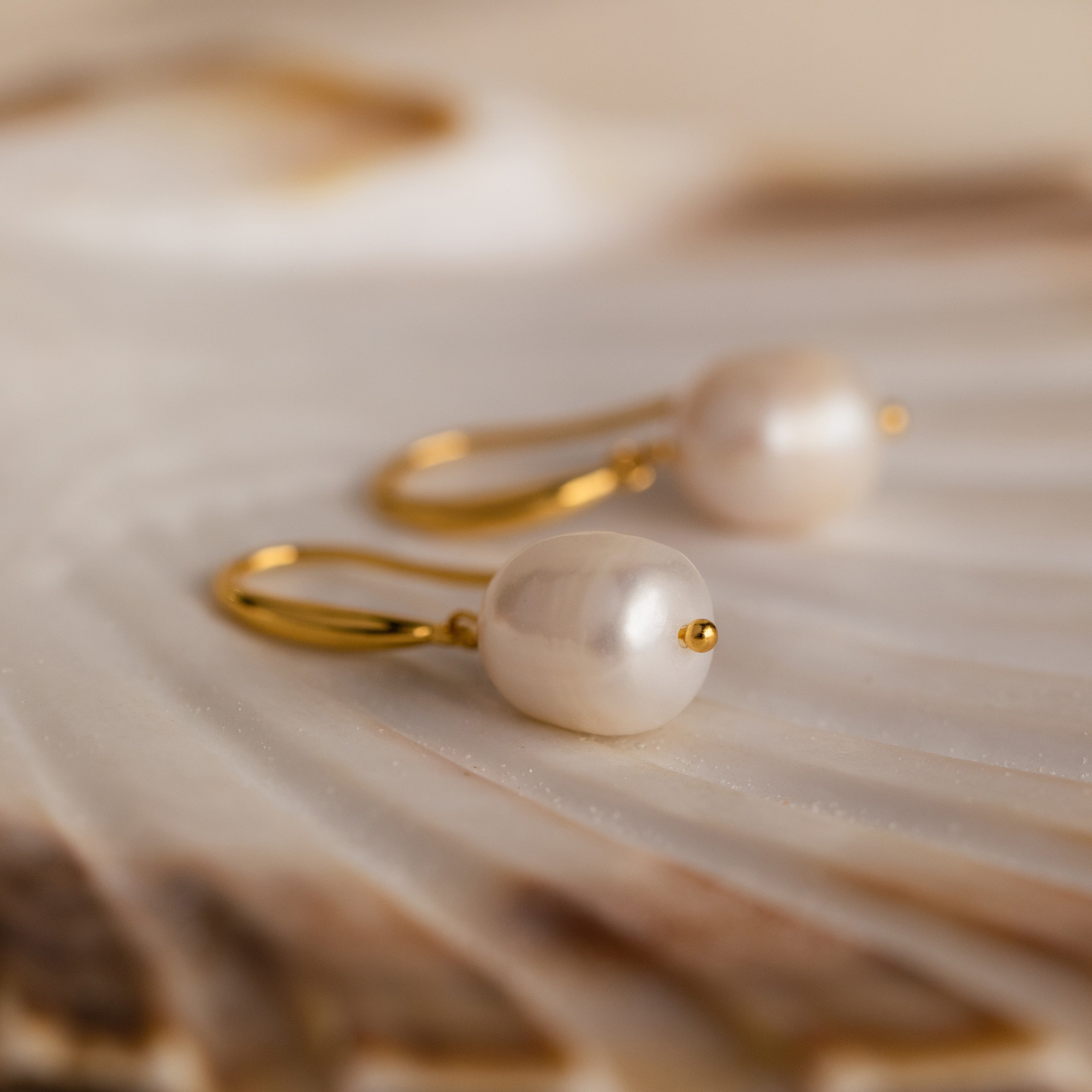 A pair of gold Dangling Pearl Drop Earrings resting on a shell-like textured surface.