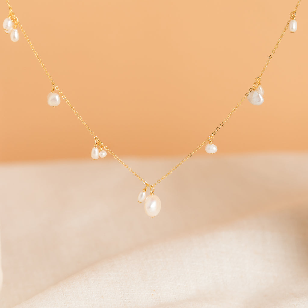 The Baroque Pearl Station Necklace in 18K Gold features a delicate gold chain with small pearls, shown on a soft peach and beige background—an elegant choice for wedding jewelry.