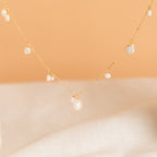 The Baroque Pearl Station Necklace in 18K Gold features a delicate gold chain with small pearls, shown on a soft peach and beige background—an elegant choice for wedding jewelry.