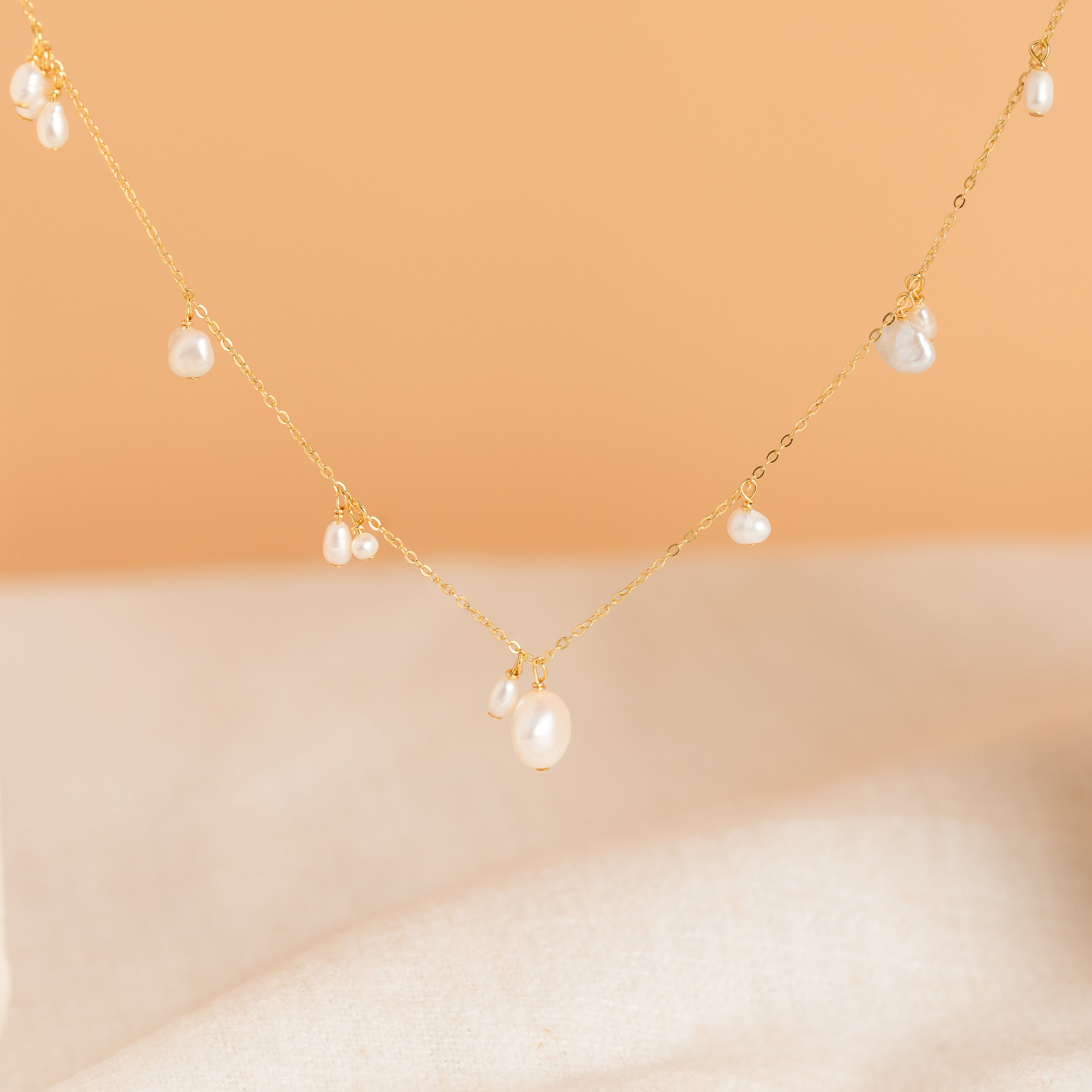 The Baroque Pearl Station Necklace in 18K Gold features a delicate gold chain with small pearls, shown on a soft peach and beige background—an elegant choice for wedding jewelry.