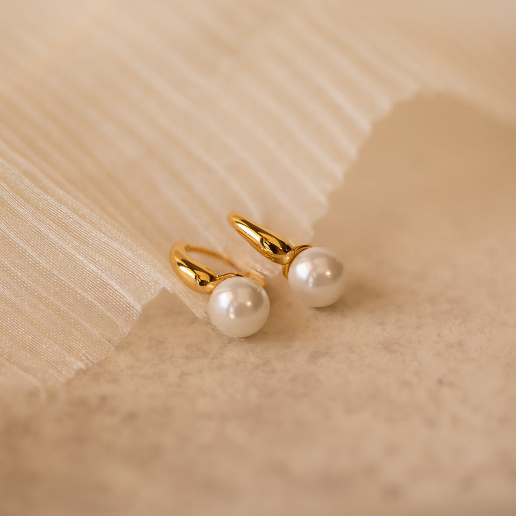 A close-up product shot of a pair of elegant gold-tone huggie earrings, each featuring a smooth, round white pearl drop.