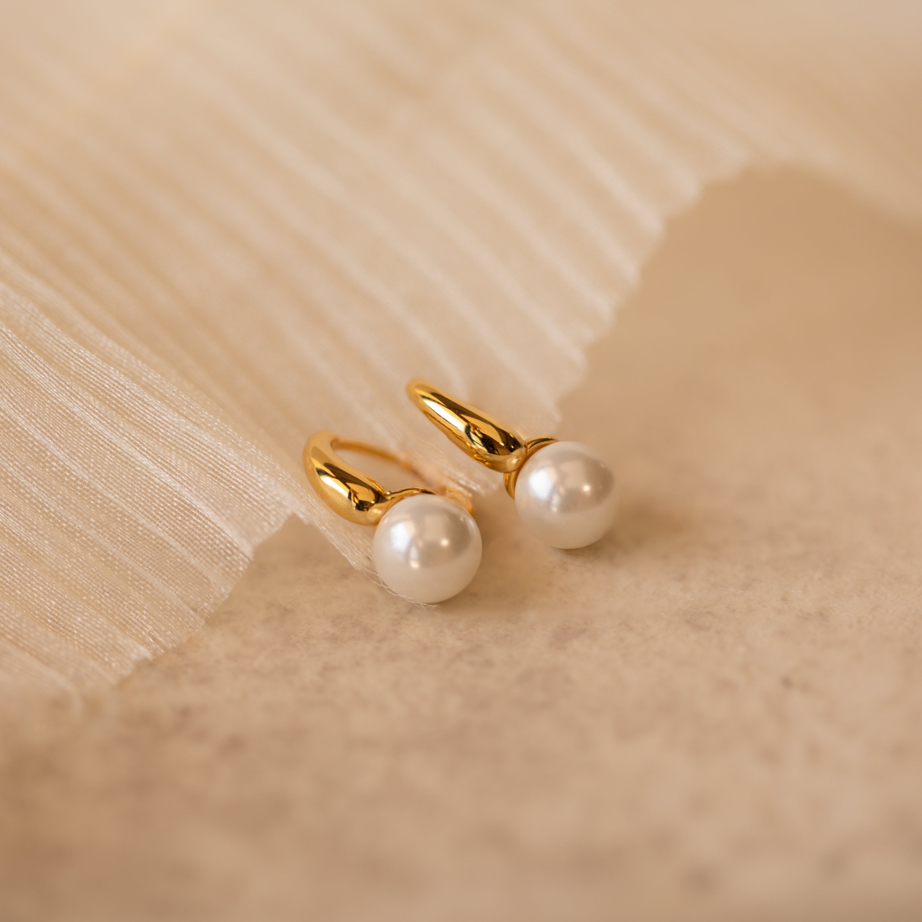 A close-up product shot of a pair of elegant gold-tone huggie earrings, each featuring a smooth, round white pearl drop.