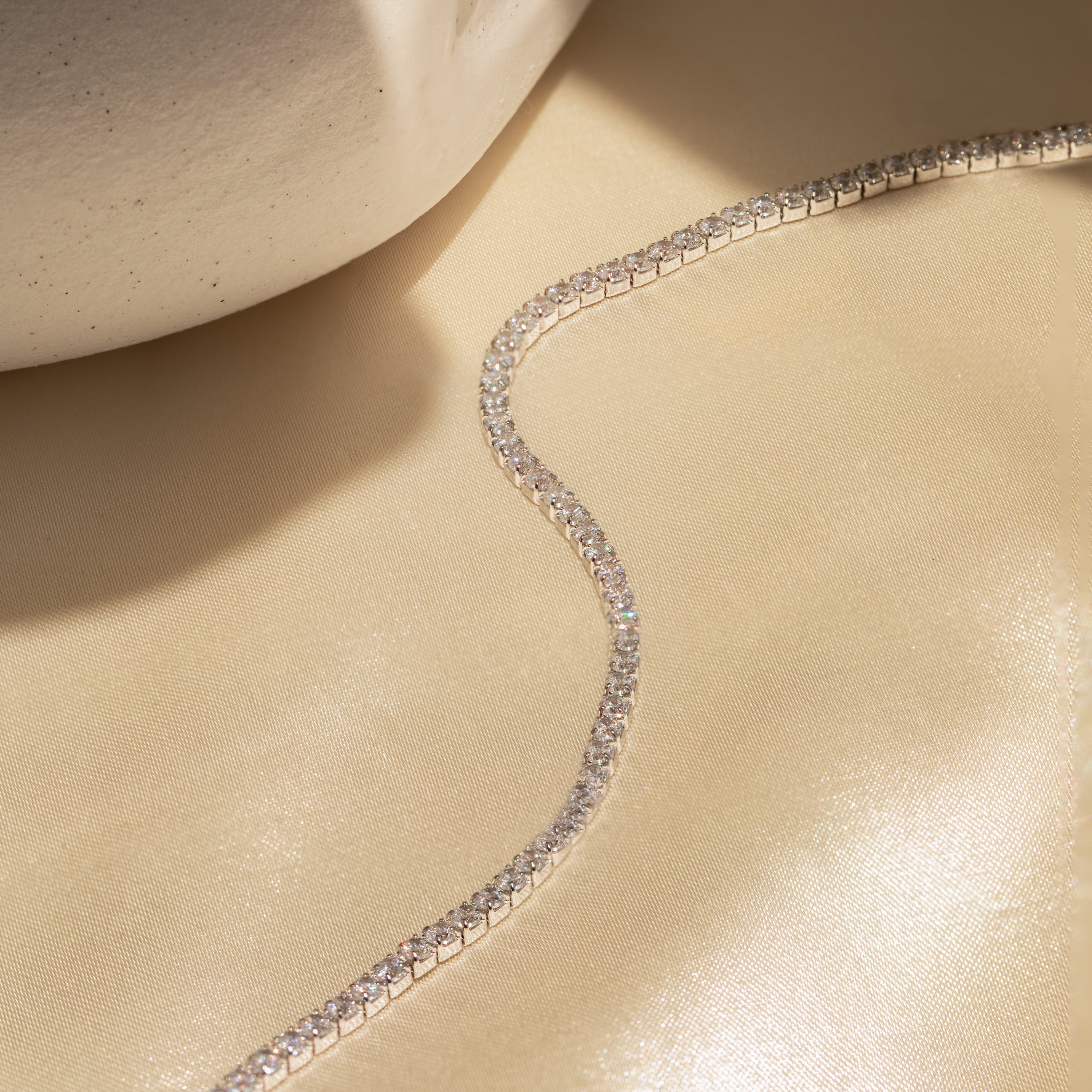 The Classic Diamond Tennis Bracelet in White Gold shines on beige fabric beside a textured ceramic piece.