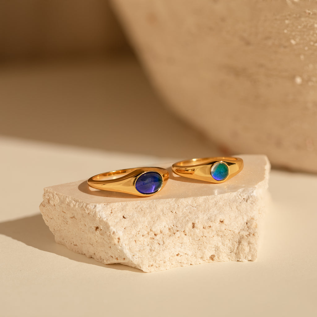 The Signet Mood Rings Set features two gold rings with blue and green stones, displayed on a cream stone slab—nostalgic, color-changing jewelry that captures mood magic.