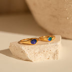 The Signet Mood Rings Set features two gold rings with blue and green stones, displayed on a cream stone slab—nostalgic, color-changing jewelry that captures mood magic.