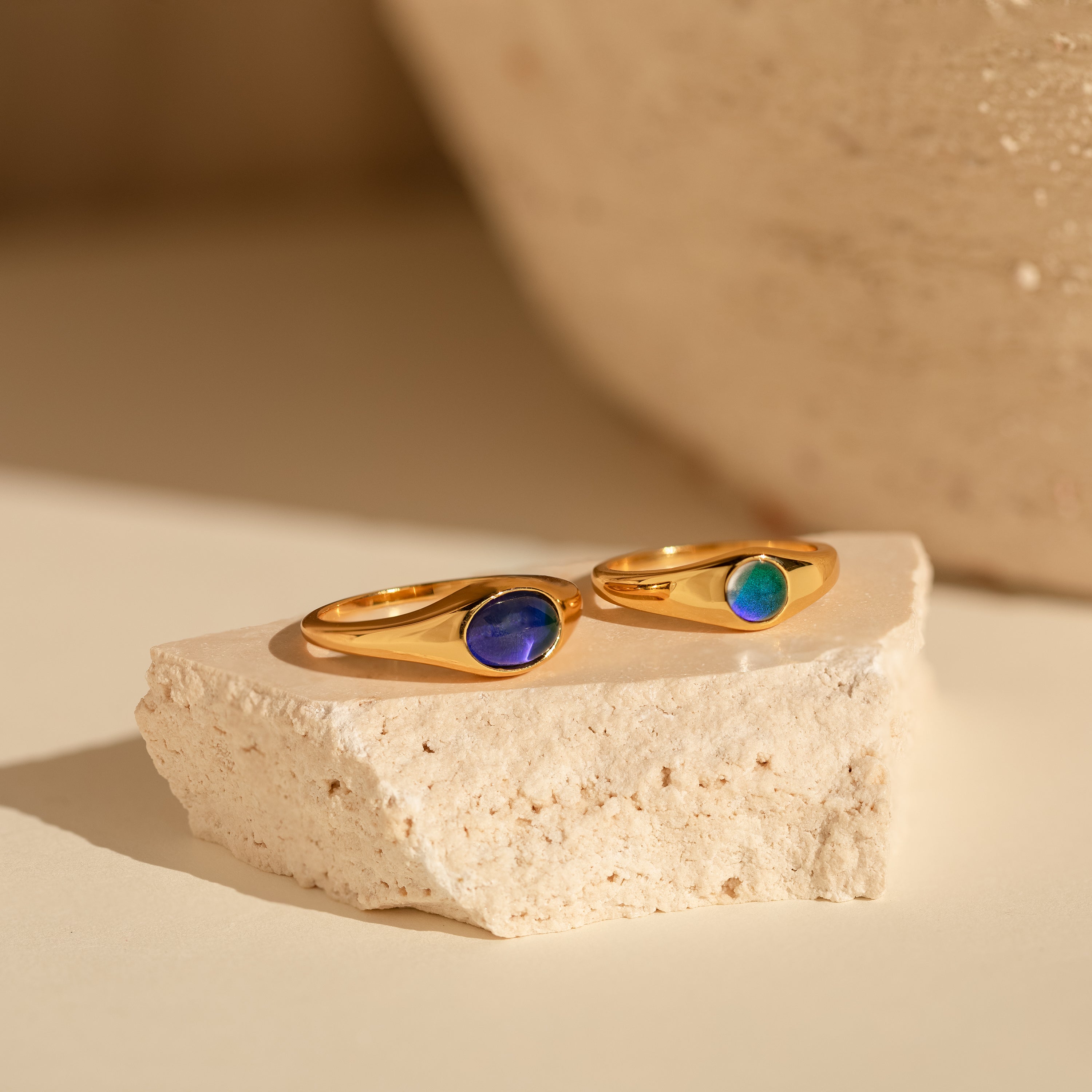 The Signet Mood Rings Set features two gold rings with blue and green stones, displayed on a cream stone slab—nostalgic, color-changing jewelry that captures mood magic.