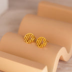 Block Monogram Earrings in round gold with a geometric cut-out design rest on a curved beige display—ideal for enhancing your earring stack.