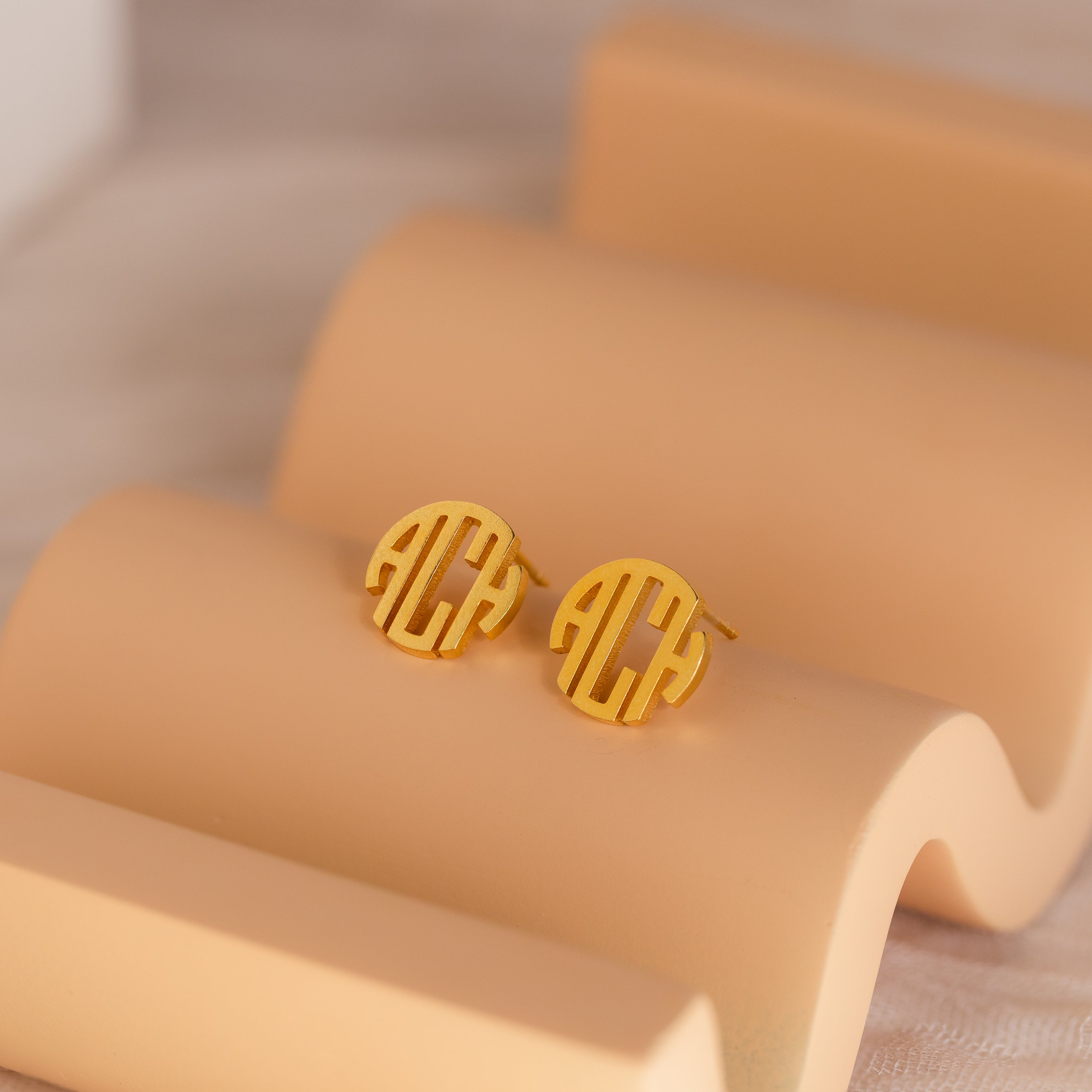 Block Monogram Earrings in round gold with a geometric cut-out design rest on a curved beige display—ideal for enhancing your earring stack.
