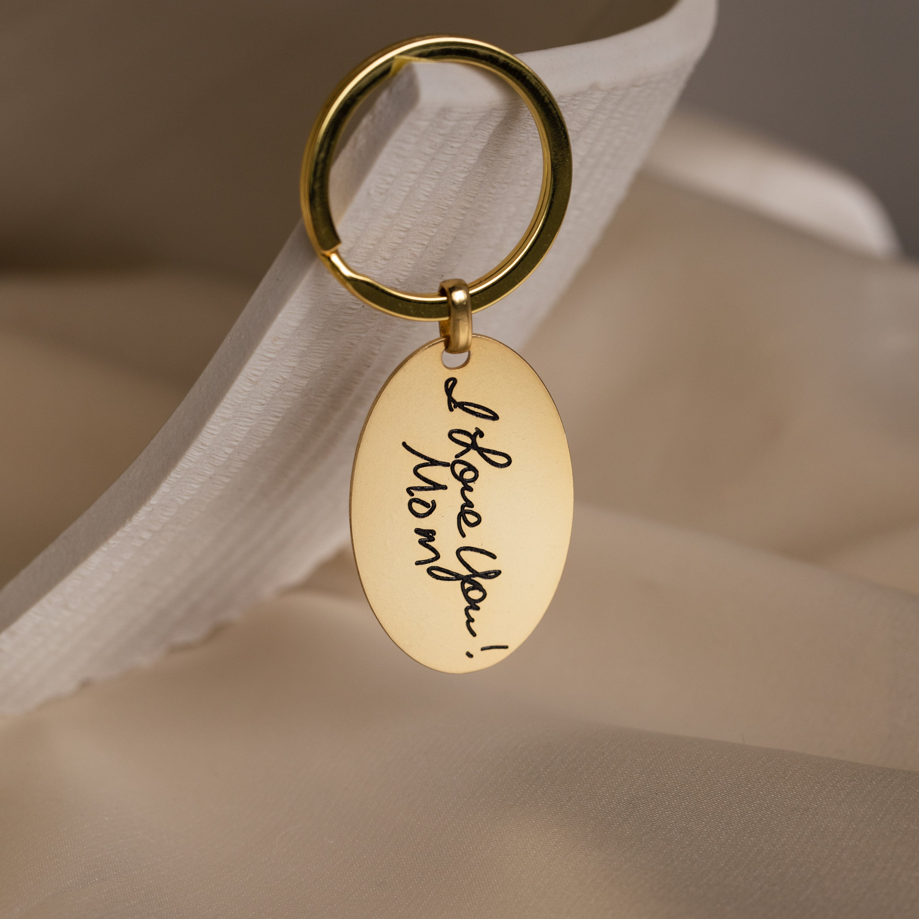 The Handwriting Oval Keychain features an oval tag engraved with "I love you! Mom" in gold script, displayed on a cream fabric background.