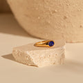 A nostalgic jewelry piece, the Signet Mood Ring in gold with a dark blue stone rests on light-colored textured stone.