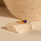 A nostalgic jewelry piece, the Signet Mood Ring in gold with a dark blue stone rests on light-colored textured stone.