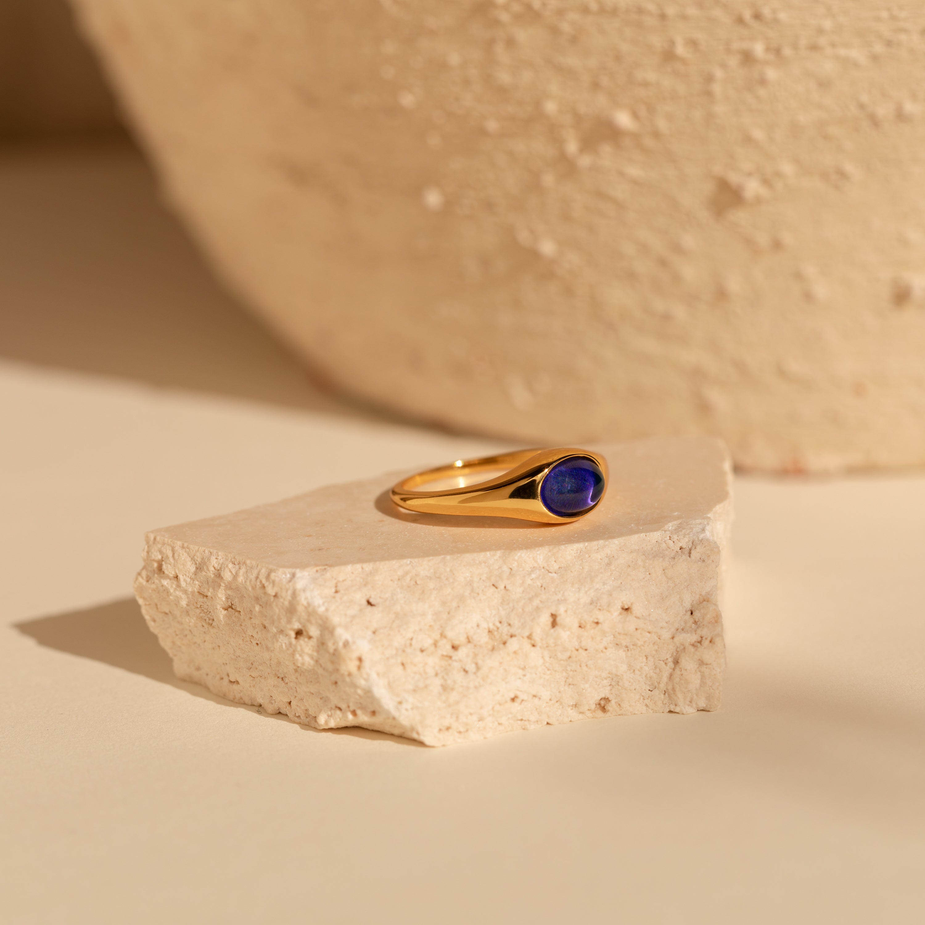 A nostalgic jewelry piece, the Signet Mood Ring in gold with a dark blue stone rests on light-colored textured stone.