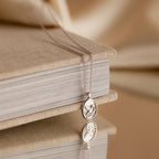 The Pearl Hummingbird Necklace in Sterling Silver is displayed draped over a closed beige book, its elegant design reflected on a glossy surface.