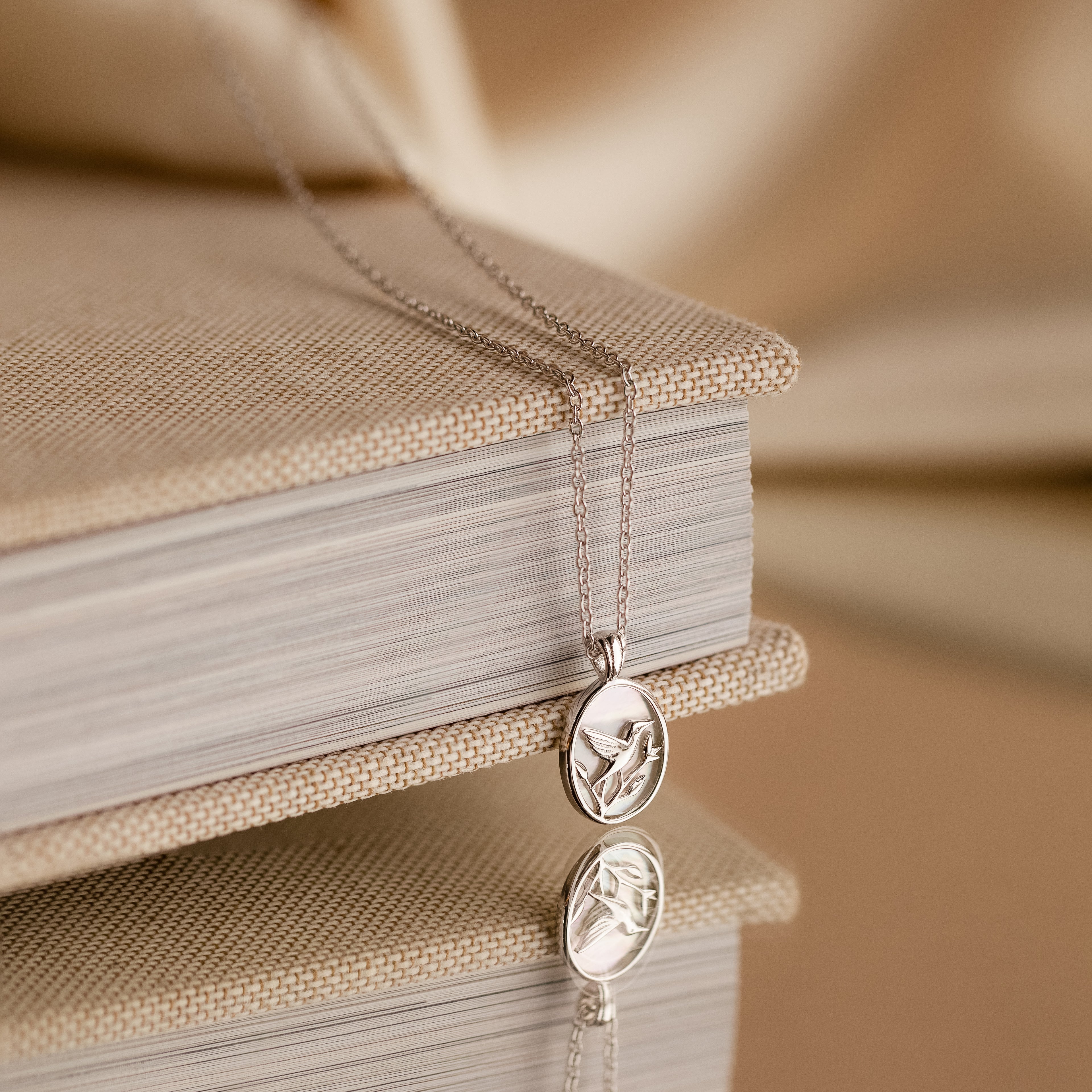 The Pearl Hummingbird Necklace in Sterling Silver is displayed draped over a closed beige book, its elegant design reflected on a glossy surface.